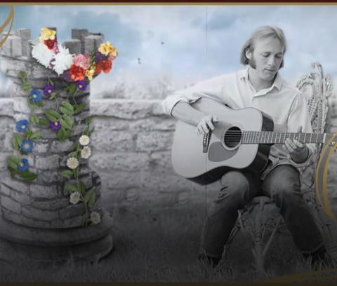 Stephen Stills sits next to a tower covered in flowers. In his lap is an acoustic guitar. This image is a screen grab from the Youtube Video