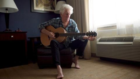 Graham Nash sits in a hotel room tuning an acoustic guitar