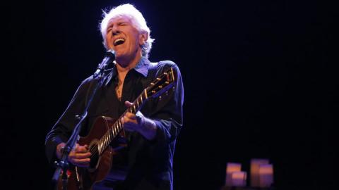 Graham Nash appears on stage in front of a microphone while playing an acoustic guitar, Graham's eyes closed and face in a deep in song